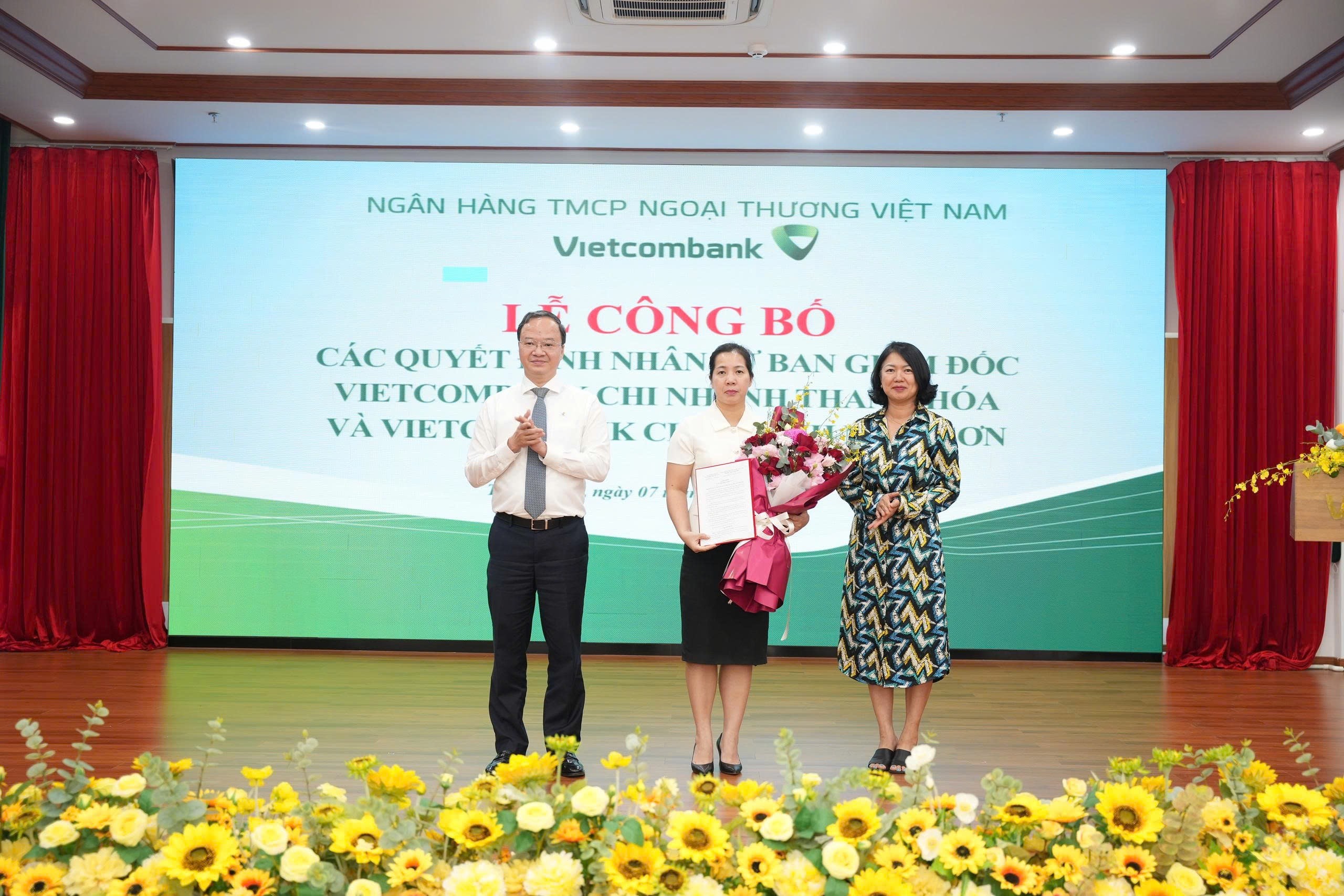 Ông Lê Quang Vinh - Phó Bí thư Đảng ủy, Tổng Giám đốc (bên trái) trao Quyết định và bà Nguyễn Thị Hồng Vân - Giám đốc Khối Pháp chế và Tuân thủ (bên phải) tặng hoa chúc mừng bà Lê Thị Hiền - tân Bí thư Chi bộ, Giám đốc Vietcombank chi nhánh Thanh Hóa