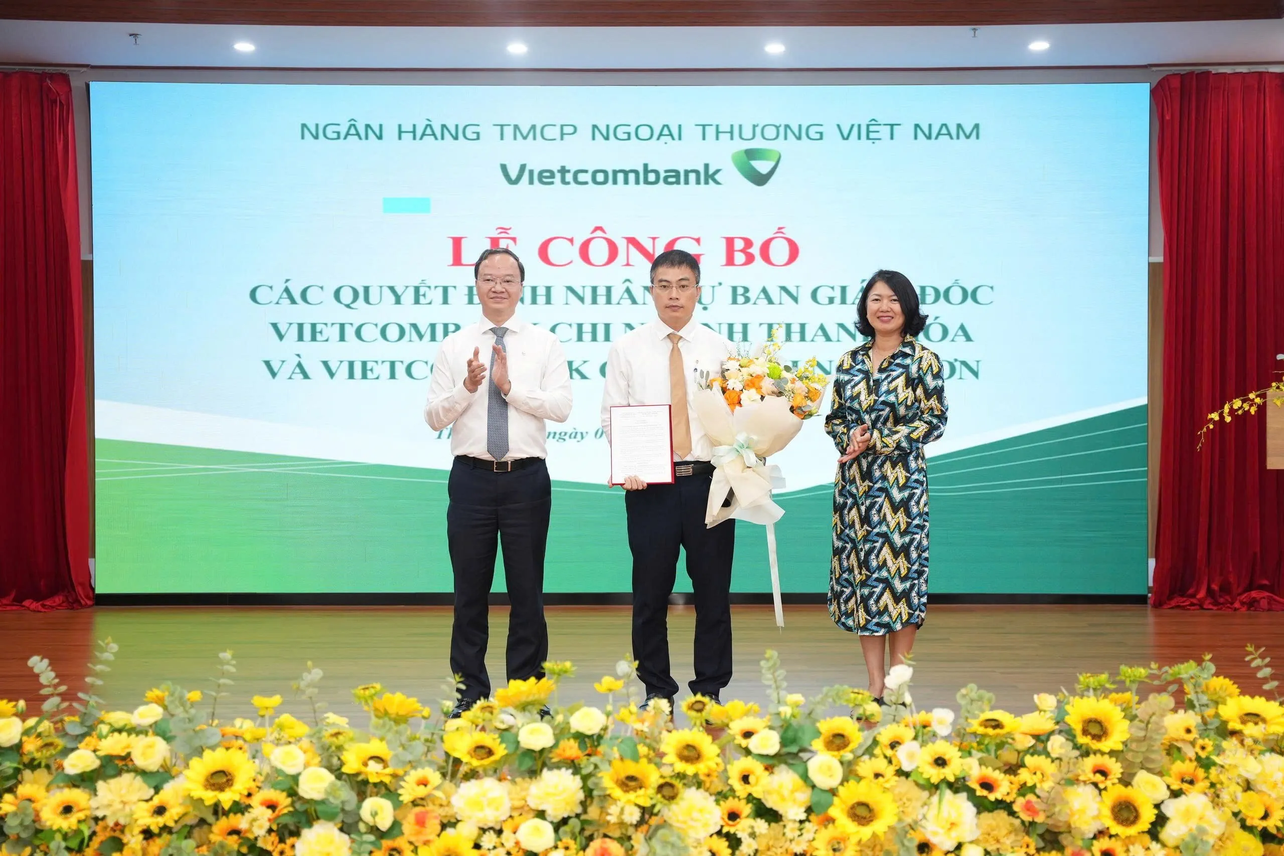 Ông Lê Quang Vinh - Phó Bí thư Đảng ủy, Tổng Giám đốc (bên trái) trao Quyết định và bà Nguyễn Thị Hồng Vân - Giám đốc Khối Pháp chế và Tuân thủ (bên phải) tặng hoa chúc mừng ông Mai Danh Tiến - tân Phó Giám đốc Vietcombank chi nhánh Nghi Sơn