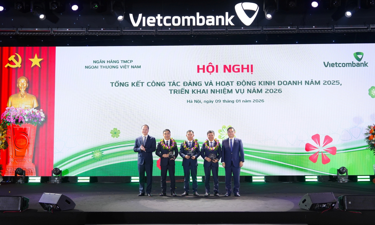 Đ/c Nguyễn Thanh Tùng - Ủy viên BCH Đảng bộ Chính phủ, Bí thư Đảng ủy, Chủ tịch HĐQT Vietcombank (ngoài cùng bên phải) và đ/c Lê Quang Vinh - Phó Bí thư Đảng ủy, Thành viên HĐQT, Tổng Giám đốc Vietcombank (ngoài cùng bên trái) trao biểu trưng và tặng hoa vinh danh cho đại diện 03 đơn vị hoàn thành đặc biệt xuất sắc nhiệm vụ kinh năm 2025 gồm Vietcombank Sở giao dịch, Vietcombank Nam Sài Gòn, Vietcombank Thành Công