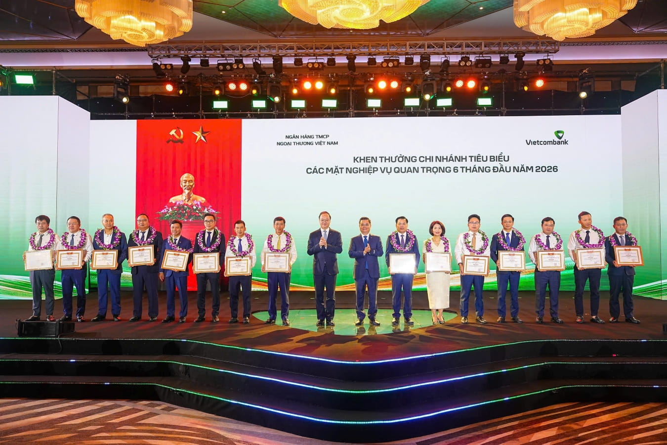 Đ/c Nguyễn Thanh Tùng - Bí thư Đảng ủy, Chủ tịch HĐQT Vietcombank (thứ 8 từ phải sang) tặng giấy khen và đ/c Lê Quang Vinh - Phó Bí thư Đảng ủy, Thành viên HĐQT, Tổng Giám đốc Vietcombank (thứ 9 từ phải sang) trao hoa chúc mừng cho 15 chi nhánh tiêu biểu trong hoạt động kinh doanh 6 tháng đầu năm 2026